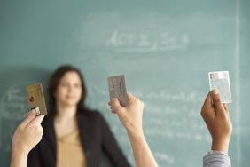 Three students raise their hands, each grasping a credit card, while in the background, a teacher standing before a blackboard looks on with a bemused expression.