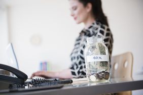 Woman working at a desk with a jar of cash labeled 401 K sitting on the desk..