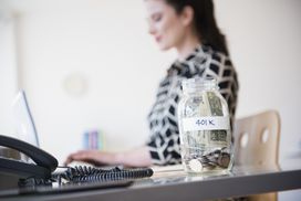 Woman working at a desk with a jar of cash labeled 401 K sitting on the desk..