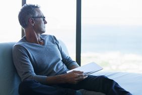 Concerned older man with tablet looking out window at expansive skyline