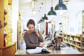 Female business owner looks at accounting paperwork while on phone at store counter 
