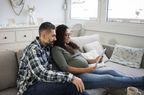 A man with a beard and a pregnant woman sit on a couch looking over their budget on a tablet