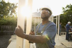 A man working on a construction site wields a drill driver as the sun rises behind him.
