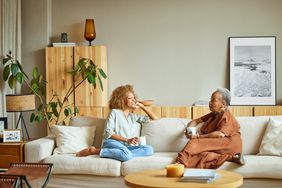A young woman and an older woman sit on a couch talking with coffee mugs in hand