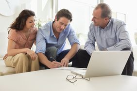 A man and a woman sit with another man while they sign a document on a table with a laptop and glasses