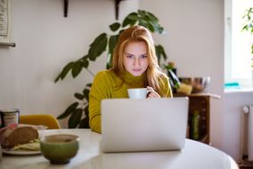 Young woman sitting at table looking at her laptop and drinking coffee. Female using laptop at home