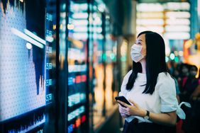 Businesswoman with protective face mask checking financial trading data on smartphone by the stock exchange market display screen board