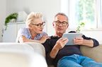 Elderly man sitting on sofa in the living room at home and showing something on digital tablet his wife.