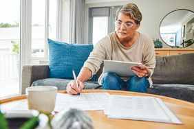 A person holds a tablet and writes on paperwork while sitting on a couch