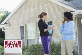 A real estate agent stands in front of a house with a for sale sign, talking with another woman