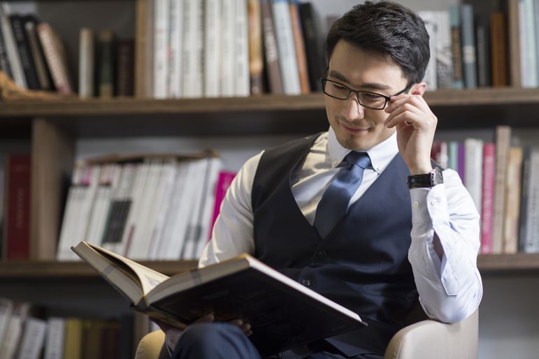 Man with glasses in a dress shirt, tie, and vest reading a book