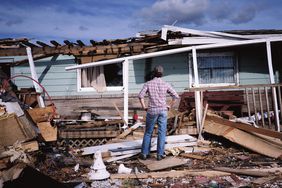 A man stands in front of a destroyed house, hands on his hips.