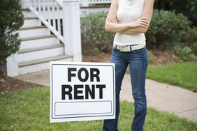 Woman standing with her arms crossed behind a "for rent" sign.