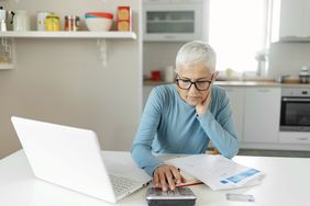 Woman in blue top with short gray hair working with a calculator and laptop in her kitchen.