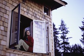 A woman in red flannel sits in the open window of a cabin and gazes out with an expression of supreme contentment.