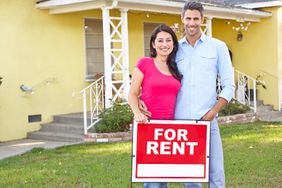A smiling couple standing outside a house with a red "for rent" sign