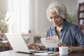Older woman at a kitchen table looking at paper work and entering into information into laptop