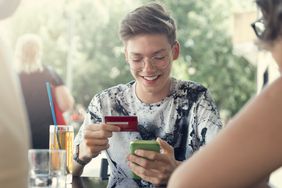 Teen boy holds his first credit card and an iphone at a table